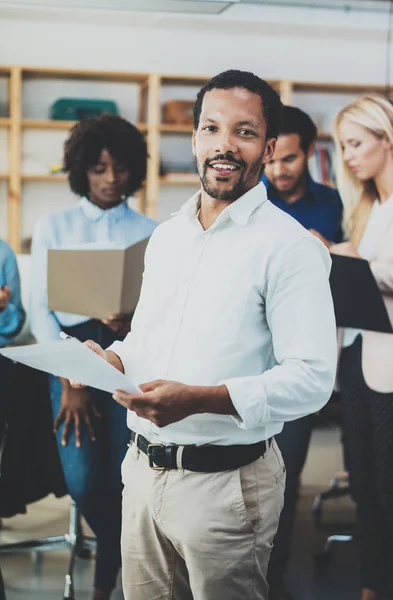 Teamwork concept in moderne kantoor. Jonge Afrikaanse zakenman dragen witte shirt houden papieren in handen en staande voorkant van het team van collega's. Verticale, onscherpe achtergrond. — Stockfoto