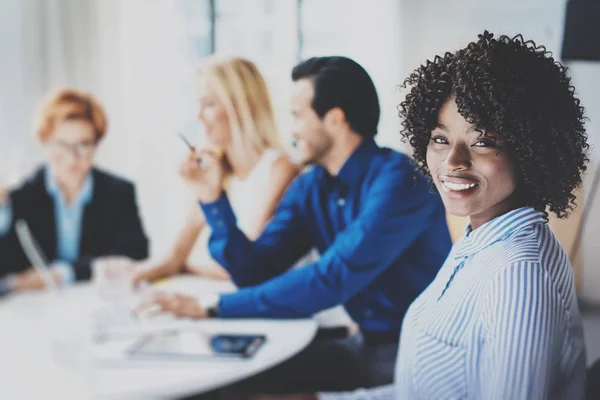 Ritratto di donna d'affari piuttosto africana americana con afro sorridente alla camera.Coworking team nel processo di brainstorming sullo sfondo in sala riunioni. Orizzontale, sfocato . — Foto Stock