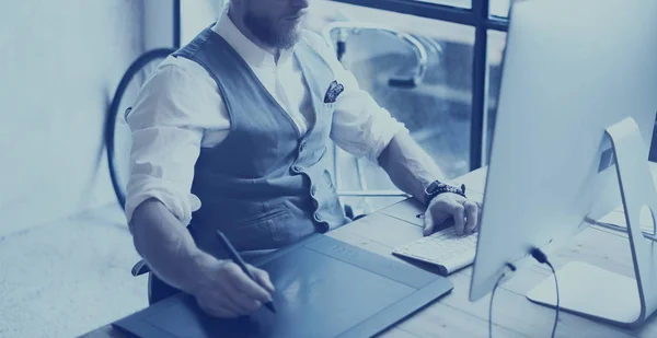Крупный план рисунка бородатого креативного менеджера с цифровой таблицей. Стильный молодой человек в белой рубашке, жилетке и работает за настольным компьютером на современном высоком уровне. Горизонтальный, киноэффект, размытый . — стоковое фото