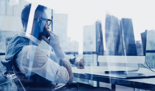 Vousatý mladý podnikatel nosit bílou košili, vestu a práce na počítači na dřevěný stůl. Koncept úspěšného podnikání. Dvojexpozicí, budova mrakodrap rozmazané pozadí. — Stock fotografie