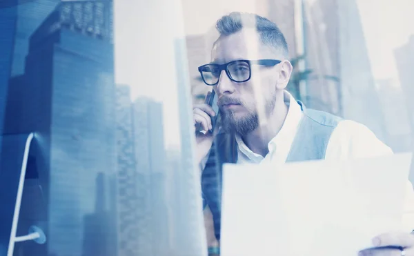 Empresario joven barbudo que usa camisa blanca, chaleco y que hace llamada en el lugar de trabajo.Concepto de negocio acertado.Exposición doble, edificio de oficina del rascacielos en fondo borroso . — Foto de Stock