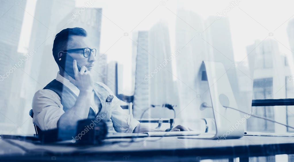 Attractive bearded young businessman wearing white shirt,waistcoat and making call at the working place.Concept of successful business.Double exposure,skyscraper office building on blurred background.