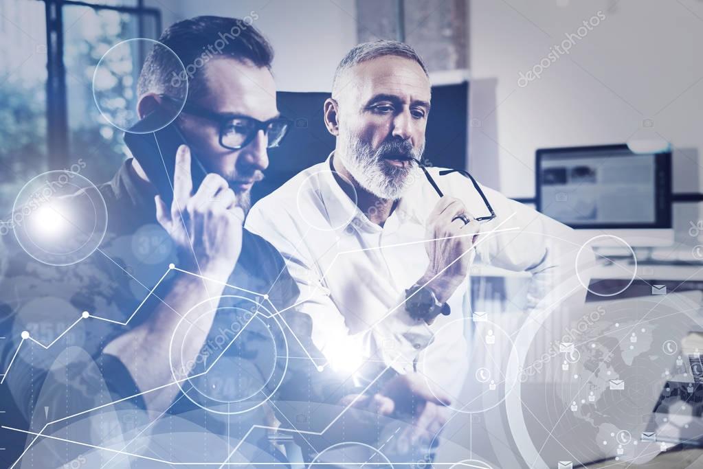 Concept of digital screen,virtual connection icon,diagram, graph interfaces.Bearded young man using mobile phone and adult colleague looking to laptop for find great work solution.Film effect,flare.