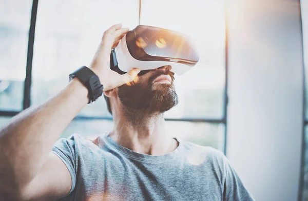 Attractive bearded man enjoyingvirtual reality glasses in modern interior design coworking studio.Home play concept.Smartphone use with VR goggles headset.Flare and sunny effect,blurred background. — Stock Photo, Image