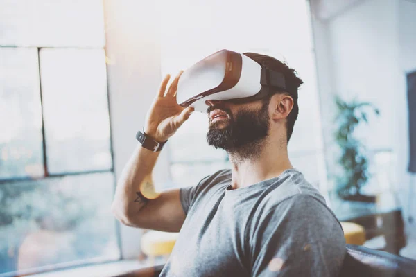 Attraktiva skäggig man med hjälp av virtuell verklighet glasögon i modern interiör coworking studio. Hipster njuter smartphone med Vr glasögon headset. Horisontell, film effekt, flare, suddig bakgrund. — Stockfoto