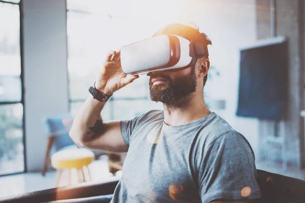 Hombre barbudo atractivo que usa gafas de realidad virtual en el interior moderno estudio de coworking. Hipster usando teléfono inteligente con gafas VR auriculares. Horizontal, efecto de película, destello, fondo borroso . —  Fotos de Stock