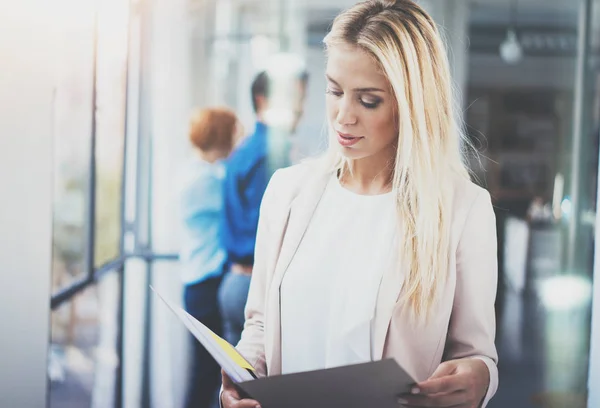 Porträtt av ung vacker blond affärskvinna i moderna coworking studio med business-team på bakgrunden. Vacker kvinna nära fönstret håller papper i händerna. Horisontell, suddig. — Stockfoto