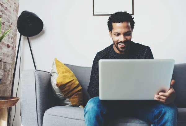 Joven hombre africano sonriendo y utilizando el ordenador portátil mientras está sentado en su casa moderna studio.Concept de personas felices working.Blurred fondo . —  Fotos de Stock