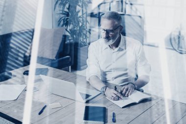 Klasik gözlüklü ve modern coworking yerde ahşap masa çalışan yetişkin başarılı işadamı kavramı. Çift pozlama, gökdelen ofis binası arka plan bulanık..