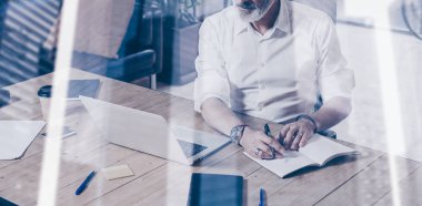 Klasik gözlüklü ve modern coworking yerde ahşap masa çalışan yetişkin başarılı işadamı kavramı. Çift pozlama, gökdelen ofis binası arka plan bulanık. Yatay geniş.
