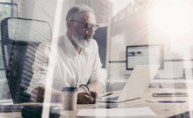 Klasik gözlüklü ve ahşap masa laptop modern coworking yerde ile çalışan yetişkin sakallı işadamı kavramı. Çift pozlama, gökdelen ofis binası arka plan bulanık..