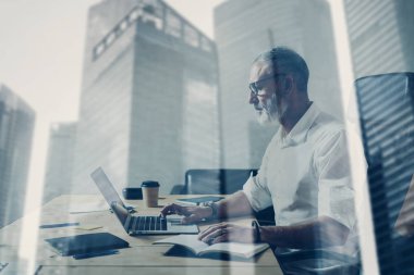 Çift pozlama kavramı. Ahşap masa modern yerde çalışan yetişkin sakallı bir iş adamı. Dizüstü bilgisayar kullanan şık orta yaş adam. Gökdelen ofis binası üzerinde arka plan bulanık. Yatay.