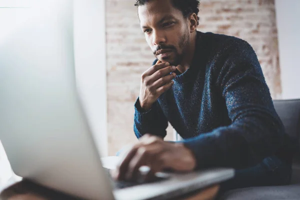 Hombre africano joven usando el ordenador portátil mientras está sentado en su moderno lugar de coworking. Concepto de la gente de negocios concentración completa. Pared de ladrillo sobre fondo borroso . —  Fotos de Stock