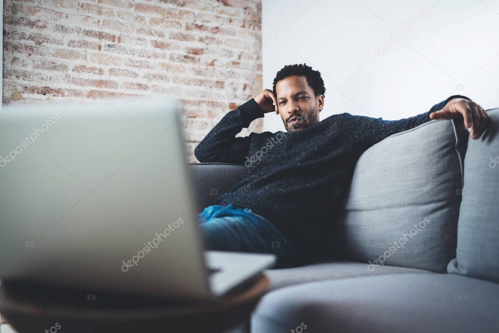 Young African confident businessman sitting on sofa at his modern coworking office and making video call via laptop.Concept of serious business people.Blurred background.