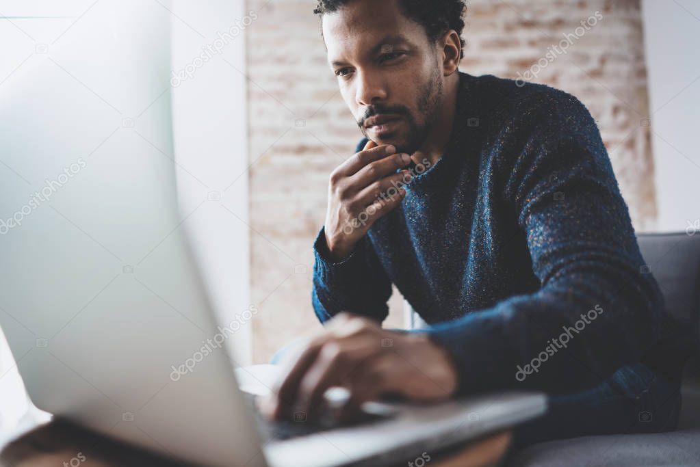 Young African man using laptop while sitting at his modern coworking place.Concept of business people full concentration.Brick wall on blurred background.