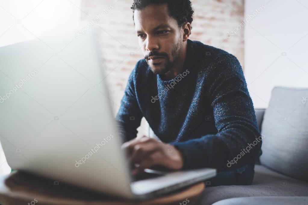 Attractive young African man using laptop while sitting at his modern coworking place.Concept of business people full concentration.Brick wall on blurred background.
