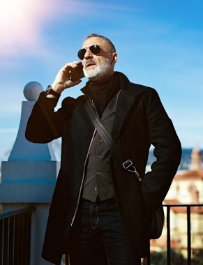 Üzerinde siyah ceket ve Şehir Parkı içinde vakit sırasında cep telefonu konuşurken ciddi çekici bir adam. Dikey, bulanık arka plan, parlama efekti.