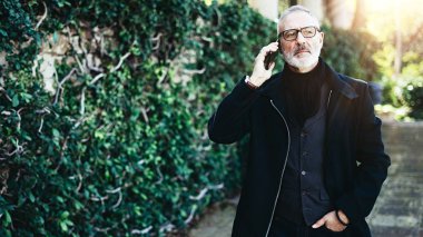 Onun Smartphone'da şehir parkında yürürken konuşan çekici yetişkin adam portresi. Yatay, bulanık arka plan, parlama efekti.
