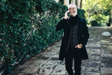 Onun Smartphone'da şehir parkında yürürken konuşmak başarılı orta yaş işadamı portresi. Yatay, bulanık arka plan, film etkisi.