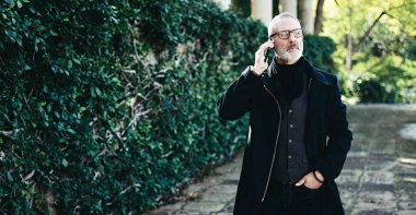 Onun Smartphone'da şehir parkında yürürken konuşmak başarılı orta yaş işadamı portresi. Yatay geniş, bulanık arka plan, film etkisi.