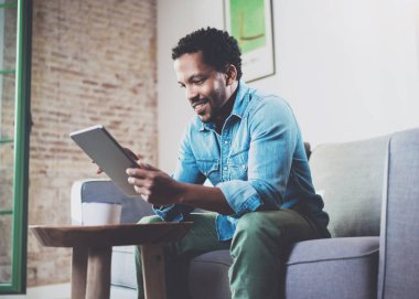 Çekici gülümseyen evde kanepede otururken çalışan Afrika erkeğin sakallı. Dijital tablet için görüntülü konuşma kullanma. Mobil cihazlar ile genç insanlar iş kavramı. Arka plan bulanık.