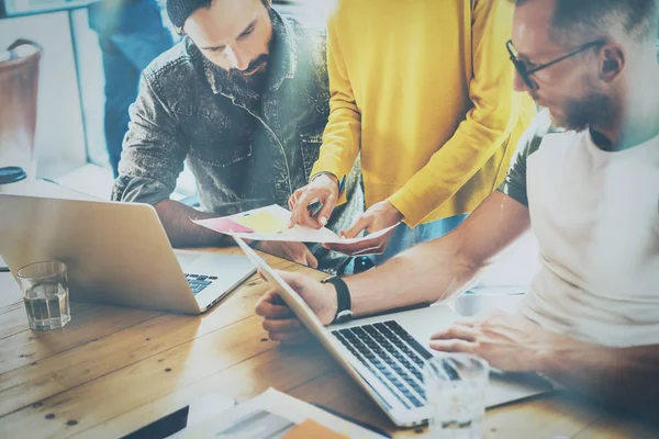 Coworking-Prozess in einem sonnigen Büro. Zwei bärtige Männer arbeiten am Computer am Holztisch. Frau trägt gelben Pullover und zeigt Dokument in der Hand.Horizontaler, verschwommener Hintergrund — Stockfoto