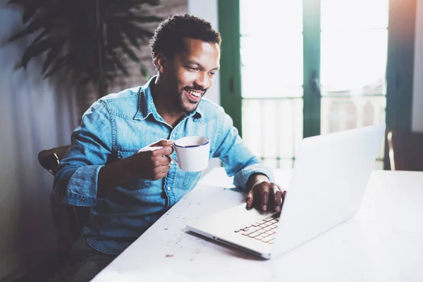 Sourire barbu homme africain travaillant à la maison tout en étant assis sur la table en bois.Utilisation d'un ordinateur portable moderne pour la recherche d'un nouvel emploi.Concept de jeunes gens travaillent appareils mobiles. . — Photo