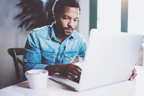 Pensativo barbudo africano que trabaja en casa mientras está sentado la mesa de madera.El uso de la computadora portátil moderna para la nueva búsqueda de empleo.Concepto de los jóvenes trabajan dispositivos móviles.Fondo borroso.Cultivo . —  Fotos de Stock