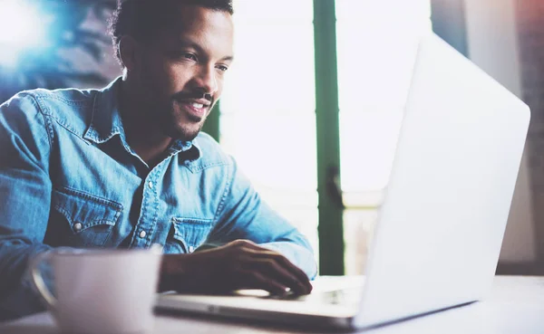 Hombre africano barbudo feliz usando el ordenador portátil en casa mientras está sentado en la mesa de madera.El tipo está escribiendo en el teclado del cuaderno. Concepto de los jóvenes trabajan dispositivos móviles. . —  Fotos de Stock