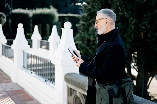 Atractivo hombre de negocios de pelo gris mensaje de texto en el teléfono móvil, mientras que pasar tiempo en el parque de la ciudad en el día soleado.Horizontal, fondo borroso . — Foto de Stock