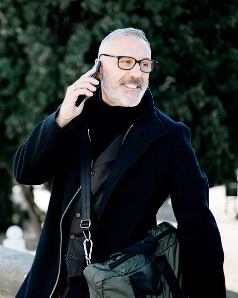 Retrato de hombre de pelo gris sonriente haciendo una llamada en su teléfono móvil mientras pasa tiempo en el parque de la ciudad en un día soleado.Fondo vertical, borroso . —  Fotos de Stock