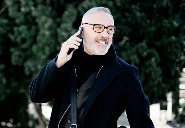 Porträtt av leende gråhårige mannen gör samtal på sin mobiltelefon medan spendera tid i stadsparken på solig dag. Horisontell, suddig bakgrund. — Stockfoto