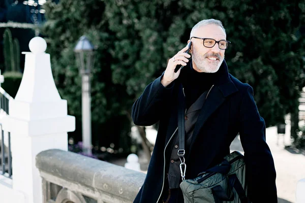 Sorrindo homem de cabelos grisalhos chamando em seu telefone celular enquanto passa o tempo no parque da cidade em dia ensolarado.Horizontal, fundo borrado . — Fotografia de Stock