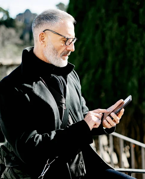 Porträt eines bärtigen, grauhaarigen erwachsenen Geschäftsmannes, der am sonnigen Tag im Stadtpark Nachrichten per Handy verschickt. Vertikaler, unscharfer Hintergrund. — Stockfoto