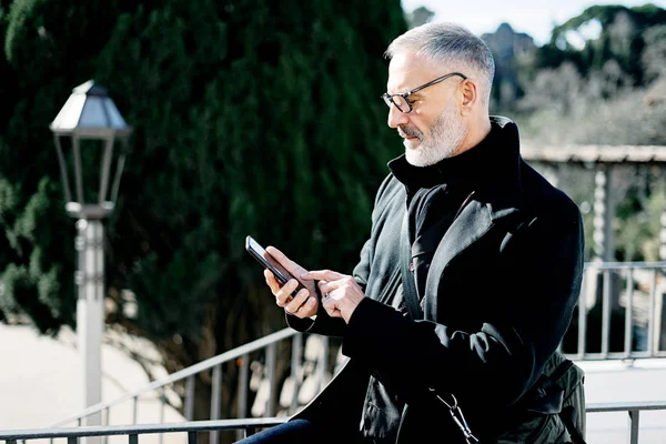 Brodaty siwy biznesmen dorosły, wysyłając wiadomość na telefon komórkowy podczas odpoczynku w parku miejskim w słoneczny dzień. Poziome, niewyraźne tło. — Zdjęcie stockowe