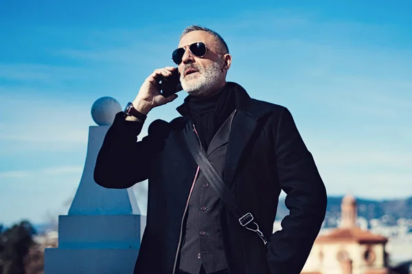 Hombre atractivo serio que usa gafas de sol de aviador negro y habla en el teléfono móvil mientras pasa tiempo en el parque de la ciudad.Fondo horizontal y borroso . —  Fotos de Stock