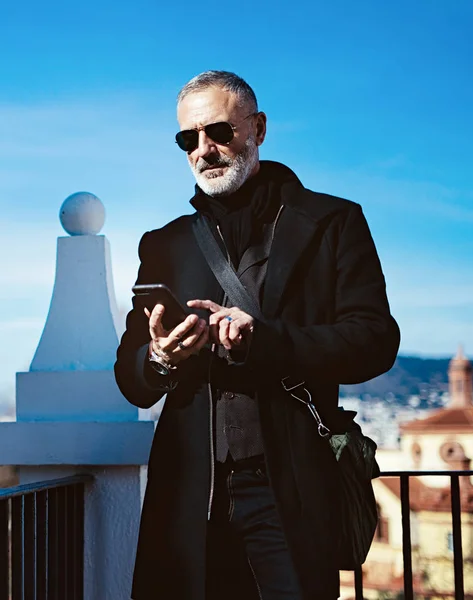 Fundersam vuxen man bär svart aviator solglasögon och trycka smartphone skärmen medan spendera tid i stadsparken. Vertikal, suddig bakgrund. — Stockfoto