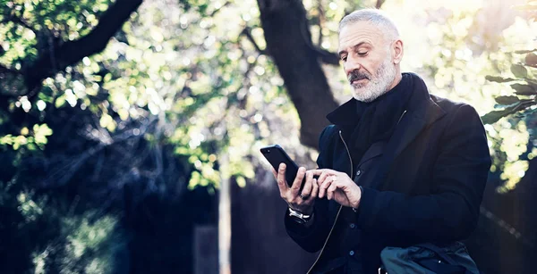 Retrato de un exitoso hombre de negocios de mediana edad usando un teléfono inteligente moderno mientras pasa tiempo en el parque de la ciudad en un día soleado. . — Foto de Stock