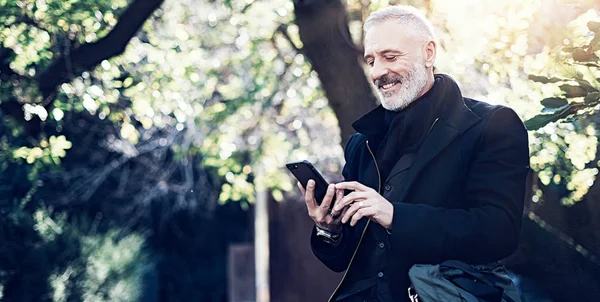 Ritratto di un uomo d'affari sorridente di mezza età che utilizza uno smartphone moderno mentre trascorre del tempo nel parco cittadino nelle giornate di sole. . — Foto Stock