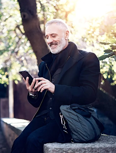 Retrato de un hombre de negocios sonriente y feliz de mediana edad usando un teléfono inteligente moderno mientras pasa tiempo en el parque de la ciudad en un día soleado. . — Foto de Stock