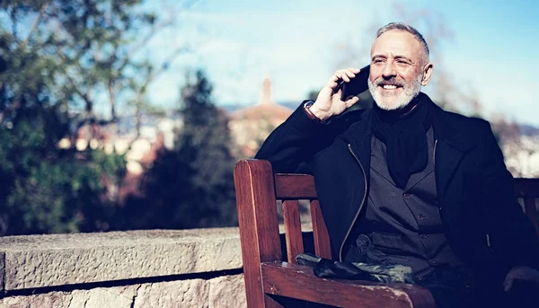 Szczęśliwy średnim wieku biznesmen w klasycznym garniturze i rozmowy na telefon komórkowy siedząc w parku miejskim. Poziome, niewyraźne tło. — Zdjęcie stockowe