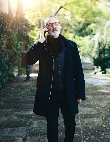 Retrato del hombre de negocios barbudo de mediana edad que habla en su teléfono inteligente mientras camina en el parque de la ciudad.Horizontal, fondo borroso, efecto de bengala . —  Fotos de Stock