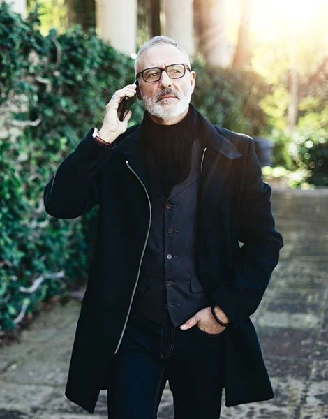 Retrato de un atractivo hombre de negocios adulto hablando en su teléfono inteligente mientras camina en el parque de la ciudad.Fondo vertical, borroso, efecto de bengala . —  Fotos de Stock