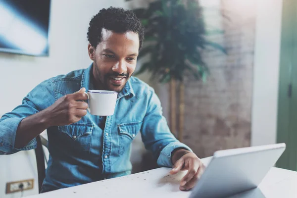 Sonriente joven africano haciendo conversación de vídeo a través de tabletas digitales con amigos mientras bebe café negro en la oficina soleada.Concepto de gente feliz de negocios.Fondo borroso, efecto de película . —  Fotos de Stock