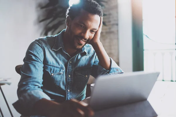 Hombre africano joven feliz haciendo conversación de videollamada a través de tableta digital con socios de negocios mientras está sentado en la oficina moderna. Fondo borroso, efecto de bengala . —  Fotos de Stock