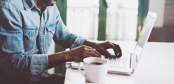 Lachende bebaarde Afrikaanse man met behulp van laptop thuis, terwijl hij aan de houten tafel zit.Man handen typen op het toetsenbord notebook.Concept van jongeren werken mobiele apparaten.Wazig venster achtergrond, breed — Stockfoto