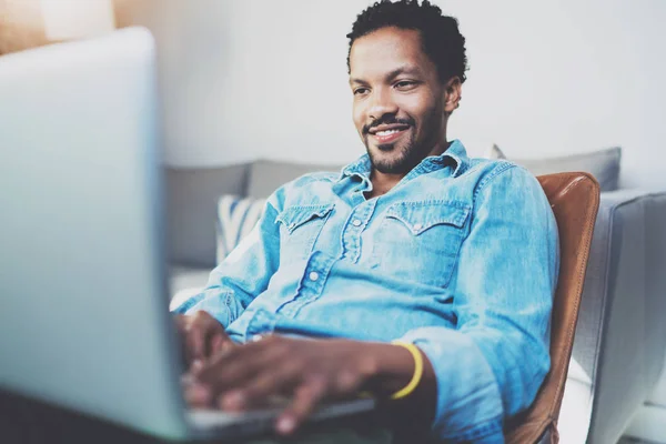 Sourire barbu homme africain faisant conversation vidéo via ordinateur portable avec des amis tout en étant assis sur un fauteuil à sa maison ensoleillée.Concept de gens d'affaires heureux.Fond flou, effet de film . — Photo