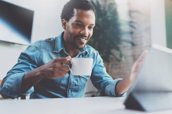 Sonriente joven africano hombre haciendo video conversación a través de tableta digital con la familia mientras bebe café negro en la oficina soleada.Concepto de gente feliz de negocios.Fondo borroso, efecto de película . —  Fotos de Stock