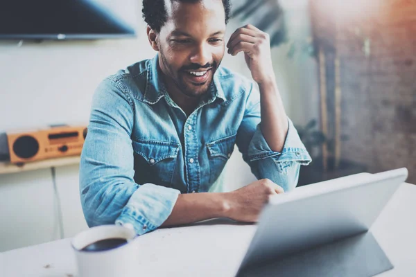 Sonriente joven africano hombre haciendo video conversación a través de tableta digital con socios de negocios mientras bebe café negro en la oficina soleada.Concepto de feliz coworking people.Blurred fondo, bengala . —  Fotos de Stock