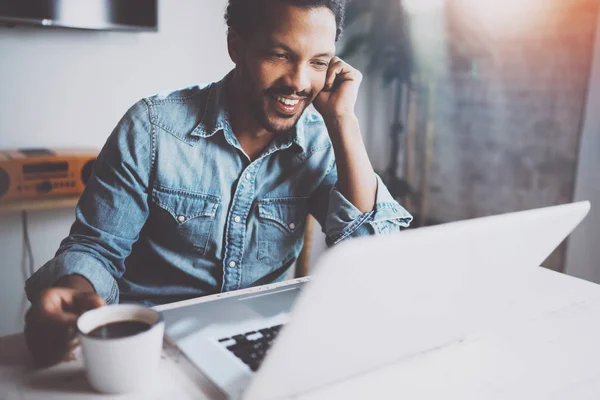 Heureux homme africain barbu faisant conversation vidéo via ordinateur portable moderne avec des partenaires tout en tenant café noir tasse blanche à la maison.Concept de jeunes gens d'affaires.Fond flou, effet fusées éclairantes . — Photo